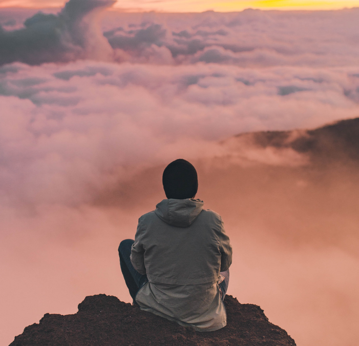 A guy sitting on a cliff looking out into the sunset